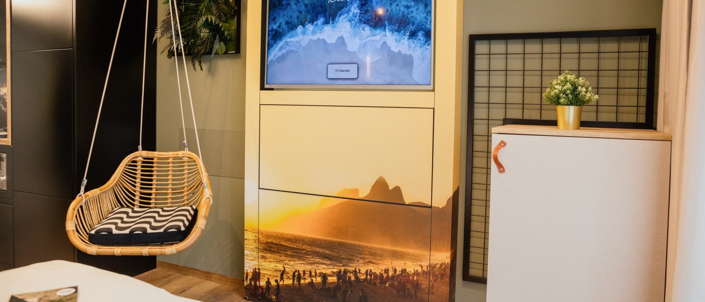 A cozy room with a hanging chair, a TV with a beach picture and a small cupboard with a plant., © Rioca Stuttgart Posto 4 GmbH A cozy room with a hanging chair, a TV with a beach picture and a small cupboard with a plant., © Rioca Stuttgart Posto 4 GmbH