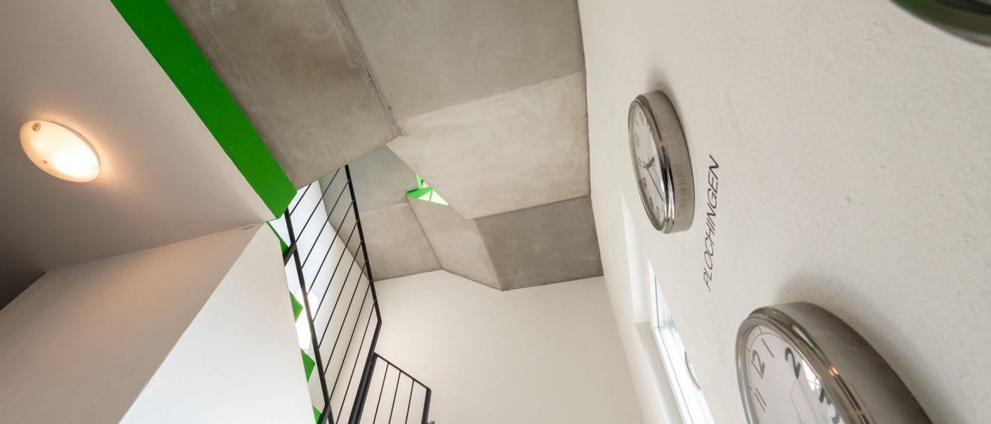 Blick in ein modernes Treppenhaus mit grünen Akzenten und mehreren Wanduhren. Die Architektur wirkt minimalistisch und funktional., © a2 HOTELS Blick in ein modernes Treppenhaus mit grünen Akzenten und mehreren Wanduhren. Die Architektur wirkt minimalistisch und funktional., © a2 HOTELS