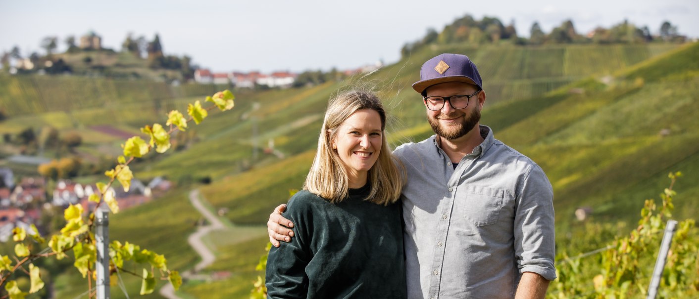 Ein Paar steht lächelnd in einem Weinberg. Im Hintergrund sind grüne Hügel und ein Dorf zu sehen. Der Mann trägt eine Mütze, die Frau hat blonde Haare., © Weinbau Wieder Ein Paar steht lächelnd in einem Weinberg. Im Hintergrund sind grüne Hügel und ein Dorf zu sehen. Der Mann trägt eine Mütze, die Frau hat blonde Haare., © Weinbau Wieder