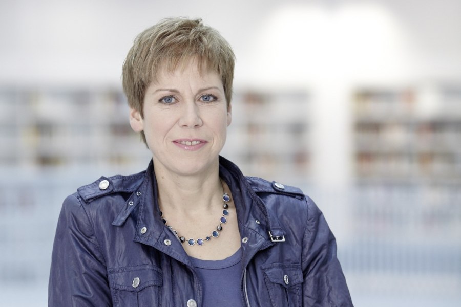 Frau mit kurzem Haar und blauer Jacke vor unscharfem Hintergrund, lächelnd in die Kamera blickend., © Foto: Volker Schrank Frau mit kurzem Haar und blauer Jacke vor unscharfem Hintergrund, lächelnd in die Kamera blickend., © Foto: Volker Schrank