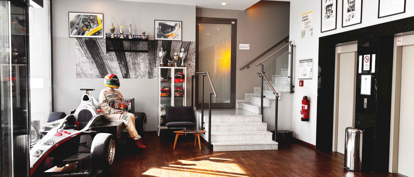 Eine moderne Lobby mit einem Rennwagen und einer Rennfahrerfigur. An den W&auml;nden h&auml;ngen Motorsportbilder und Troph&auml;en. Helle Beleuchtung und Holzboden., &copy; Novum Hospitality