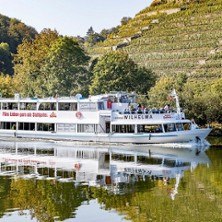 Ein Ausflugsschiff auf einem Fluss, umgeben von herbstlichen Weinbergen und Bäumen. Die Spiegelung im Wasser ist deutlich sichtbar., © links im Bild Ein Ausflugsschiff auf einem Fluss, umgeben von herbstlichen Weinbergen und Bäumen. Die Spiegelung im Wasser ist deutlich sichtbar., © links im Bild