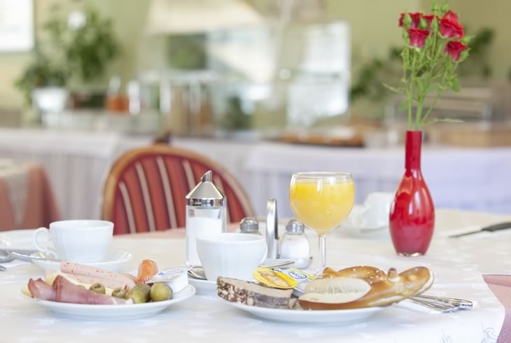 A breakfast table with rolls, cold cuts, orange juice, and a red vase of flowers in a bright room., © Hotel Drei Morgen A breakfast table with rolls, cold cuts, orange juice, and a red vase of flowers in a bright room., © Hotel Drei Morgen