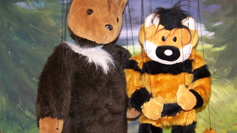 Zwei Plüschmarionetten, ein Bär und ein Tiger, hängen vor einem gemalten Waldhintergrund., © Nellys Puppentheater