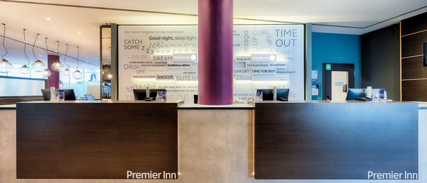Moderne Hotelrezeption mit zwei Schaltern, lila S&auml;ule und Premier Inn Logo. Im Hintergrund eine Wand mit Schlafmotiven., &copy; Premier Inn Germany