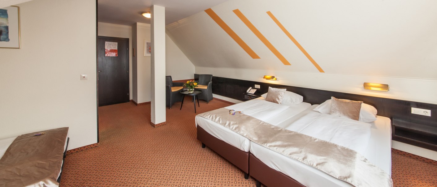 Ein gemütliches Hotelzimmer mit Doppelbett, Tisch und zwei Stühlen. Die schräge Decke ist mit orangen Streifen dekoriert. Teppichboden und dezente Beleuchtung., © Novum Management GmbH Ein gemütliches Hotelzimmer mit Doppelbett, Tisch und zwei Stühlen. Die schräge Decke ist mit orangen Streifen dekoriert. Teppichboden und dezente Beleuchtung., © Novum Management GmbH