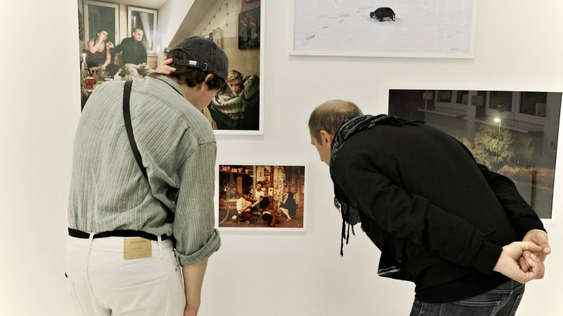 Zwei Personen betrachten aufmerksam Fotos an einer weißen Galeriewand. Die Bilder zeigen verschiedene Szenen, darunter Menschen und eine Schneelandschaft., © Palermo Galerie Zwei Personen betrachten aufmerksam Fotos an einer weißen Galeriewand. Die Bilder zeigen verschiedene Szenen, darunter Menschen und eine Schneelandschaft., © Palermo Galerie