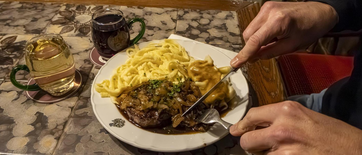 Ein Teller mit Zwiebelrostbraten, Spätzle und Soße. Daneben stehen zwei Gläser mit Getränken. Eine Person schneidet das Fleisch., © Stuttgart-Marketing GmbH, Sarah Schmid