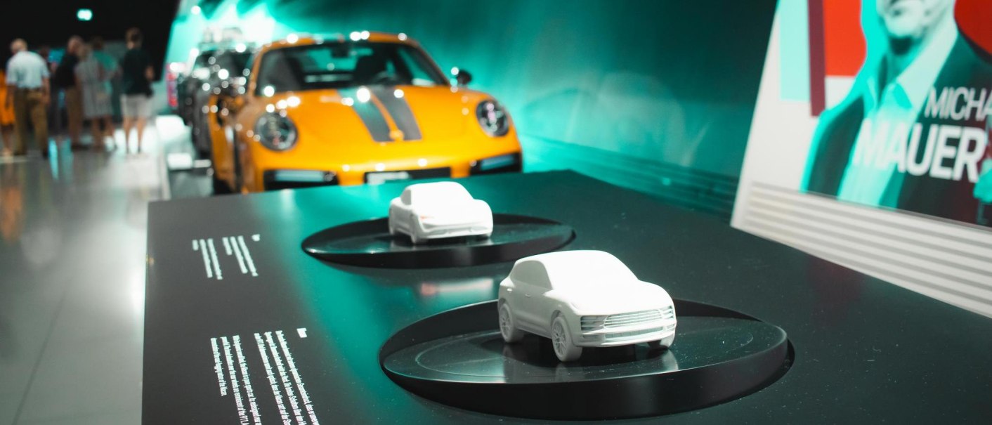 Model cars on a table in the Porsche Museum, an orange Porsche and visitors in the background. On the right, a poster with the name Michael Mauer., © Stuttgart Marketing GmbH, Sarah Schmid