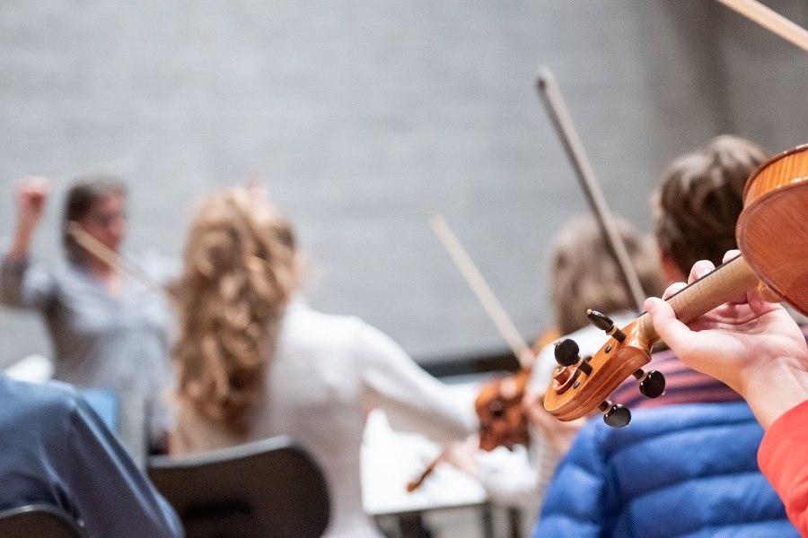 Ein Orchester probt unter der Leitung einer Dirigentin. Im Vordergrund ist eine Geige zu sehen, w&auml;hrend die Musiker konzentriert spielen., &copy; Jens Volle