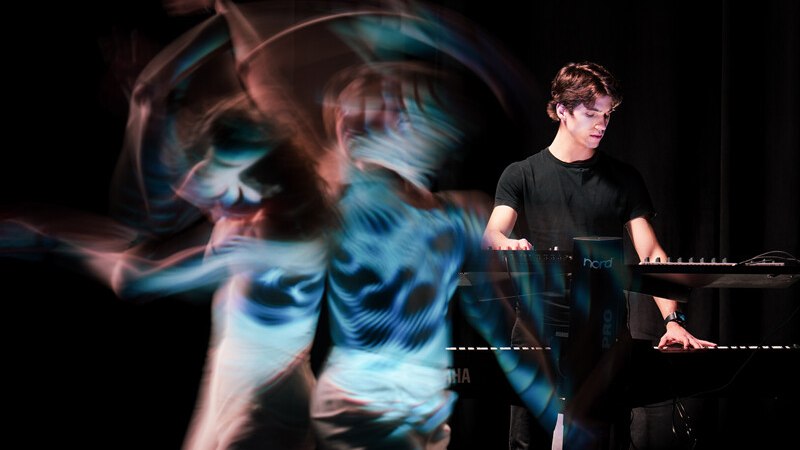 Ein Musiker spielt Keyboard, während ein Tänzer in dynamischer Bewegung verschwommen ist. Die Szene wirkt künstlerisch und energiegeladen., © Württembergische Staatstheater Stuttgart Ein Musiker spielt Keyboard, während ein Tänzer in dynamischer Bewegung verschwommen ist. Die Szene wirkt künstlerisch und energiegeladen., © Württembergische Staatstheater Stuttgart