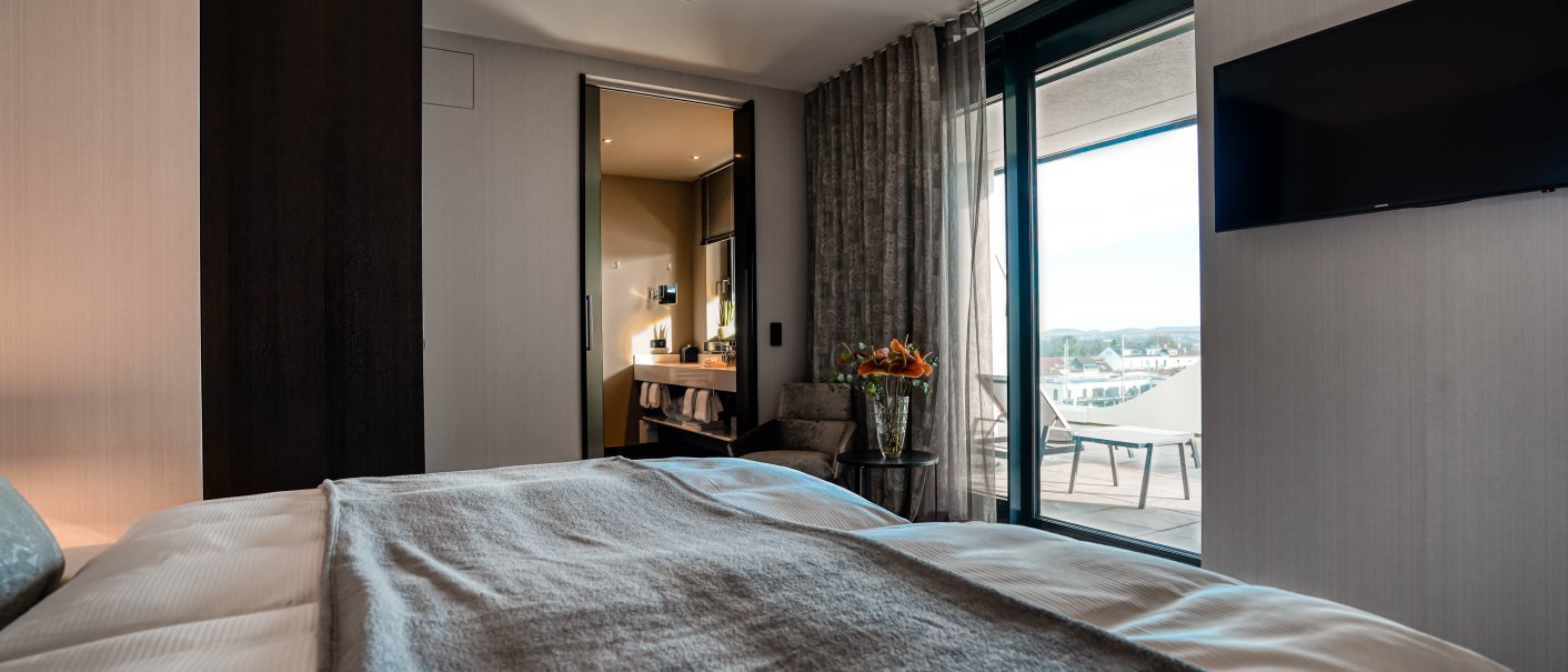 Moderne Hotelsuite mit großem Bett, Blick auf Balkon mit Sitzmöbeln. Badezimmer im Hintergrund, stilvolle Einrichtung mit Blumen und TV an der Wand., © CLOUD N°7 APARTMENTS GmbH