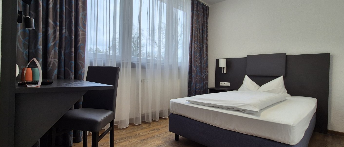 Ein schlichtes Hotelzimmer mit Einzelbett, Schreibtisch und Stuhl. Gro&szlig;e Fenster mit Vorh&auml;ngen lassen Tageslicht herein., &copy; MedientechnikKeuk