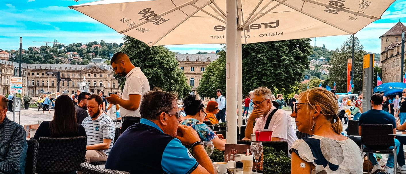 Menschen sitzen unter Sonnenschirmen in einem Café auf einer Terrasse. Im Hintergrund ist ein historisches Gebäude und eine belebte Straße zu sehen., © Café Treppe