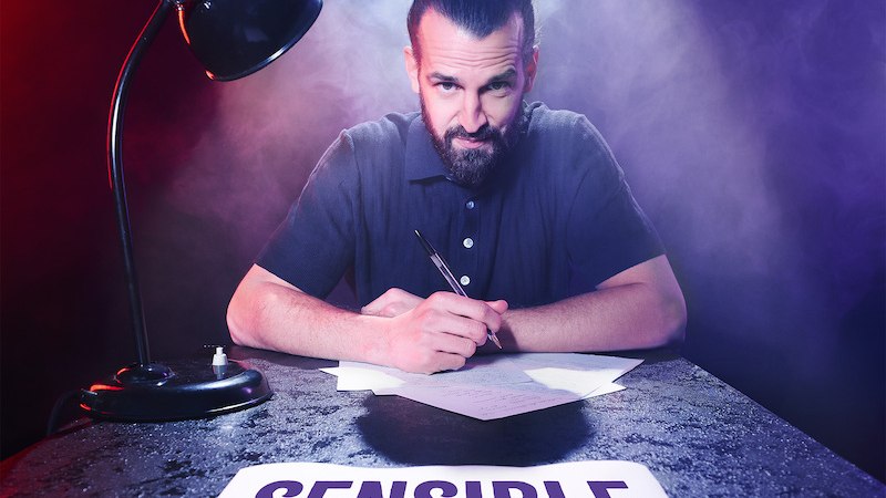 A man with a beard and a bun is sitting at a table, writing on paper. A lamp illuminates him, surrounded by red and blue light and smoke., &copy; Rosenau Kultur e.V.