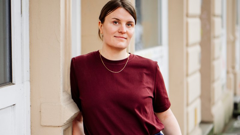 Eine Frau in einem weinroten T-Shirt lehnt entspannt an einer beigen Wand. Sie tr&auml;gt eine schlichte Halskette und blickt in die Kamera., &copy; Asja Caspari