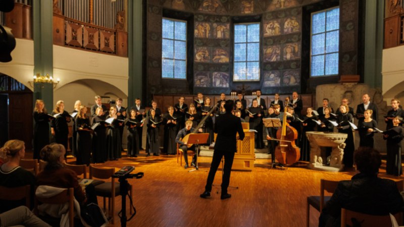 Ein Chor singt in einer Kirche, dirigiert von einer Person. Zuschauer sitzen auf Stühlen im Vordergrund und lauschen der Darbietung., © 2024 Heiko Tiedmann Ein Chor singt in einer Kirche, dirigiert von einer Person. Zuschauer sitzen auf Stühlen im Vordergrund und lauschen der Darbietung., © 2024 Heiko Tiedmann