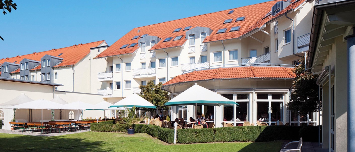 Hotel mit rotem Dach, Terrasse mit Sonnenschirmen und Sitzgelegenheiten im Garten. Sonniger Tag, blauer Himmel., © Seminaris Hotel Bad Boll