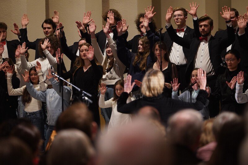 Ein Chor in formeller Kleidung singt in einer Kirche, dirigiert von einer Frau. Die S&auml;nger heben synchron die H&auml;nde, w&auml;hrend sie auftreten., &copy; Stiftsmusik Stuttgart