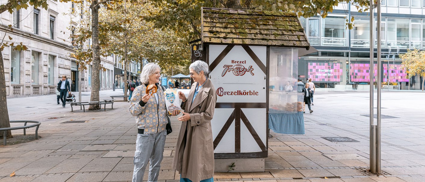Zwei Frauen lachen vor einem Brezelstand am Schlossplatz. Eine hält eine Brezel, die andere eine Tüte. Herbstliche Bäume im Hintergrund., © SMG, Sarah Schmid