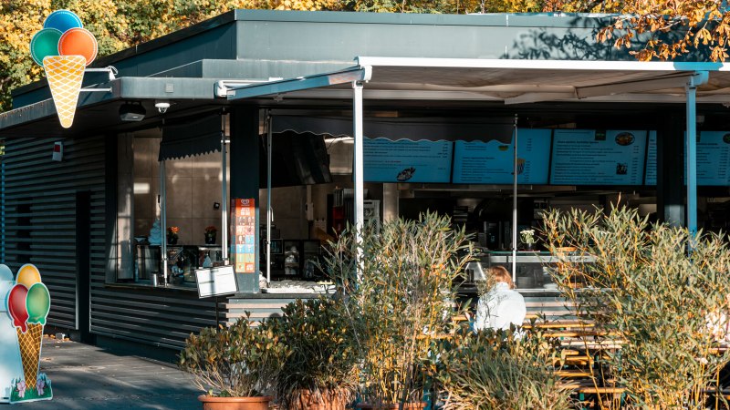 Ein Biergarten mit Eisdiele, umgeben von herbstlichen B&auml;umen. Menschen sitzen an Holztischen, w&auml;hrend ein Eis-Schild den Eingang ziert., &copy; SMG, Sarah Schmid