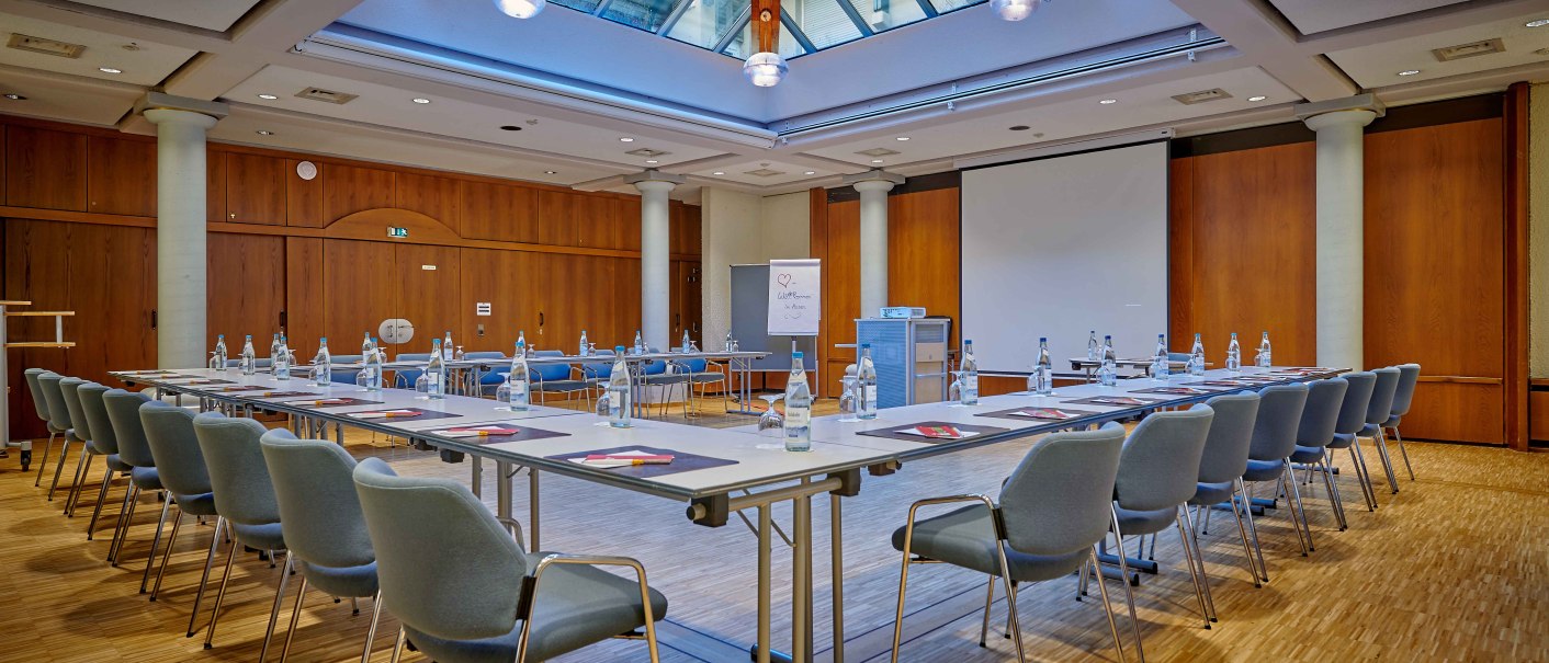 Ein Konferenzraum mit U-förmiger Tischanordnung, Stühlen, Wasserflaschen und Schreibmaterial. Ein Glasdach sorgt für natürliches Licht., © Hotel Gasthof Hasen