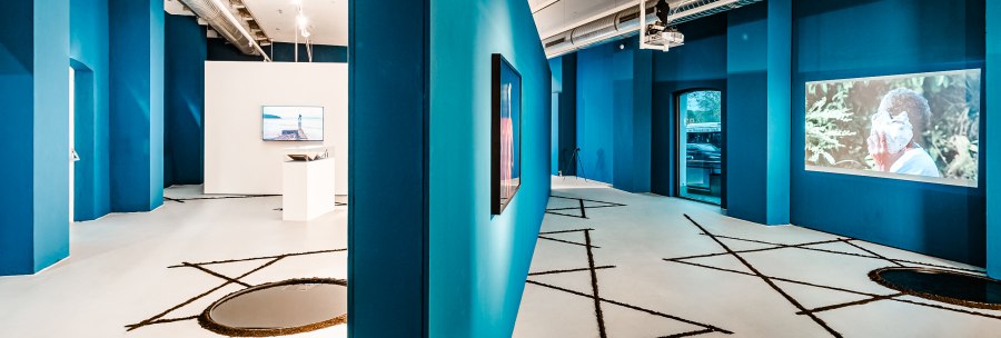 Modern gallery with blue walls, video installations and geometric patterns on the floor. Industrial design with visible ventilation pipes., &copy; Publication only by client - disclosure to third parties only after consultation.