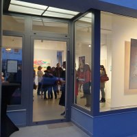Eine Galerie mit gro&szlig;en Fenstern zeigt bunte Gem&auml;lde. Menschen stehen und betrachten die Kunstwerke im Inneren., &copy; EXOgallery Contemporary and New Media Art