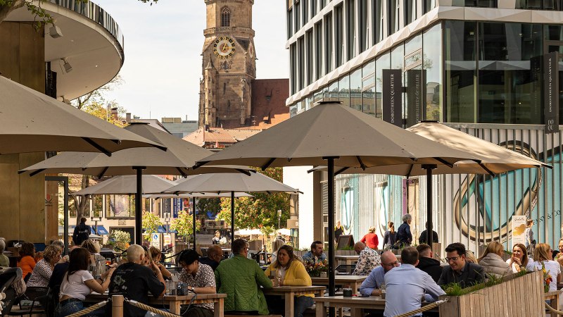 Menschen genie&szlig;en das sonnige Wetter im Dorotheenquartier in Stuttgart. Sie sitzen unter Sonnenschirmen, w&auml;hrend im Hintergrund eine Kirche zu sehen ist., &copy; Stuttgart-Marketing GmbH, Sarah Schmid