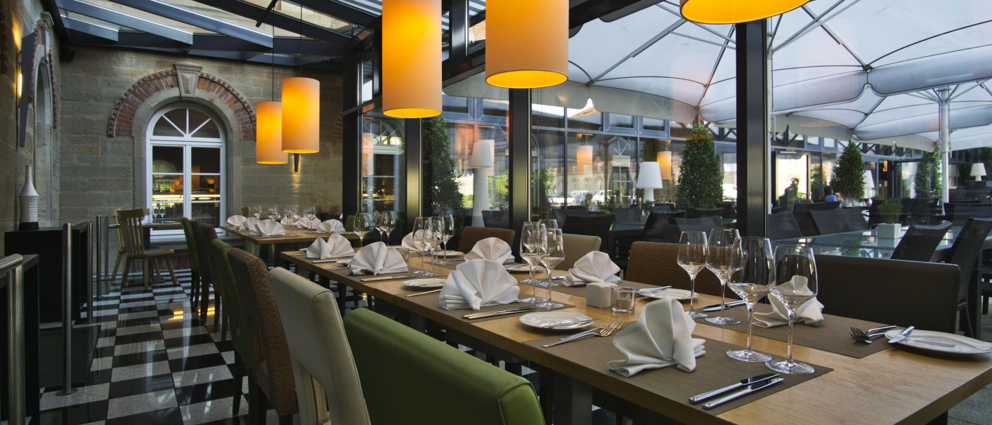 Ein elegantes Restaurant mit gedeckten Tischen, großen hängenden Lampen und einem Glasdach. Der Boden ist schwarz-weiß kariert., © TOMAS Ein elegantes Restaurant mit gedeckten Tischen, großen hängenden Lampen und einem Glasdach. Der Boden ist schwarz-weiß kariert., © TOMAS