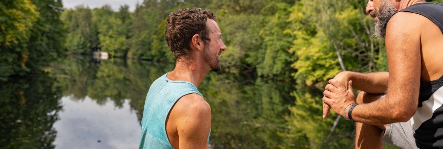 Two men sit on the shore of a lake, surrounded by green trees, and talk. One of them is holding a water bottle., &copy; SMG, Martina Denker