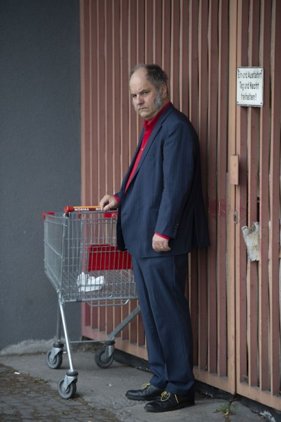 Ein Mann in einem blauen Anzug mit rotem Hemd steht ernst neben einem Einkaufswagen vor einem Holztor., &copy; Rosenau Kultur e.V.