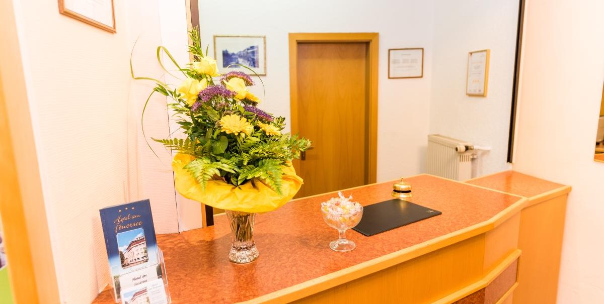 Reception at the Hotel am Feuersee with a bouquet of flowers, bell and sweets on the counter. A flyer stand is also visible., © ARNOTEL GmbH