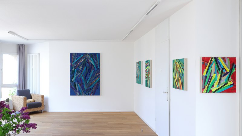 Galerie mit abstrakten Gem&auml;lden an wei&szlig;en W&auml;nden, Holzboden, Sessel und lila Blumen in einer Vase., &copy; Galerie Klaus Braun
