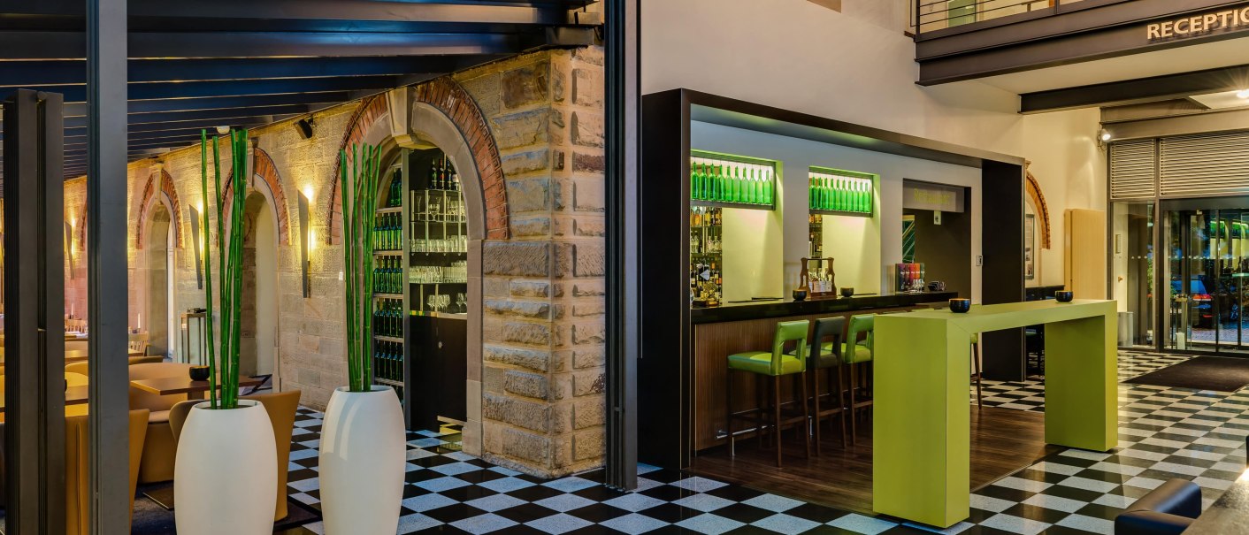 Moderne Bar mit Schachbrettboden, grünen Barhockern und hohen Pflanzen in weißen Vasen. Im Hintergrund sind Backsteinwände und eine Rezeption zu sehen., © TOMAS Moderne Bar mit Schachbrettboden, grünen Barhockern und hohen Pflanzen in weißen Vasen. Im Hintergrund sind Backsteinwände und eine Rezeption zu sehen., © TOMAS