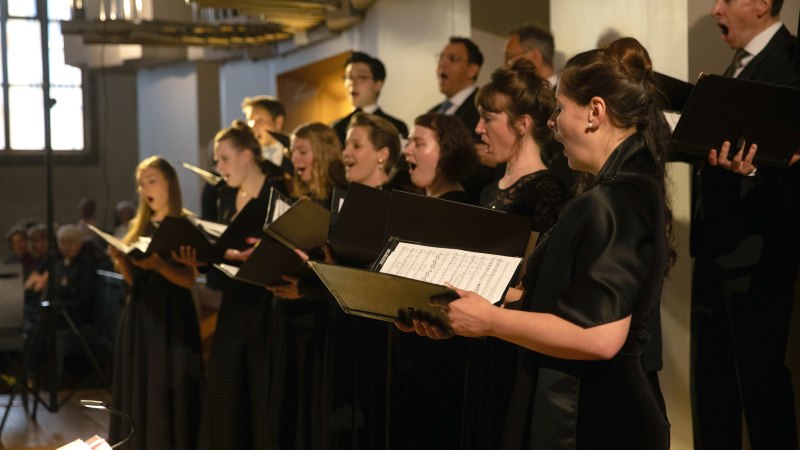 Ein Chor singt leidenschaftlich in einem Konzertsaal. Die Sänger tragen schwarze Kleidung und halten Notenblätter in der Hand., © solistenensemble stimmkunst Ein Chor singt leidenschaftlich in einem Konzertsaal. Die Sänger tragen schwarze Kleidung und halten Notenblätter in der Hand., © solistenensemble stimmkunst