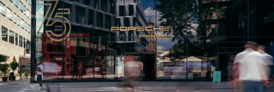 Glass façade of the Porsche Brand Store with anniversary logo '75 years 1948-2023'. People walk past, buildings and trees are reflected in the glass., © Porsche AG