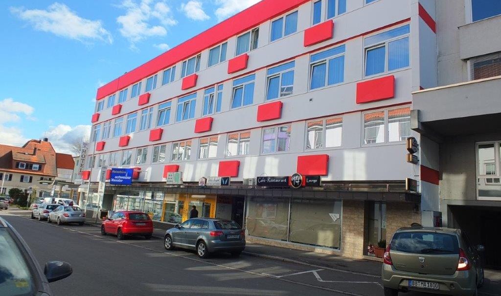 Mehrst&ouml;ckiges Geb&auml;ude mit roten Akzenten, Gesch&auml;fte im Erdgeschoss. Autos parken entlang der Stra&szlig;e, blauer Himmel mit Wolken., &copy; Hotel & Restaurant Knote