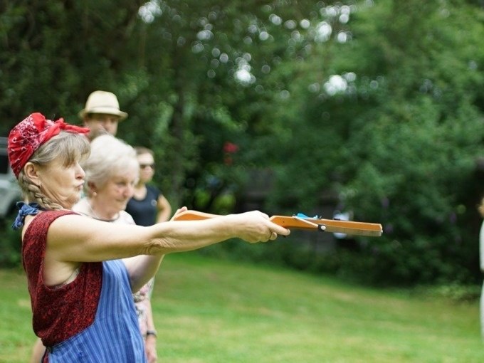 Woman with red headscarf aims wooden rifle outdoors, surrounded by other people. Green area and trees in the background., © Cool-Tours StattReisen Woman with red headscarf aims wooden rifle outdoors, surrounded by other people. Green area and trees in the background., © Cool-Tours StattReisen