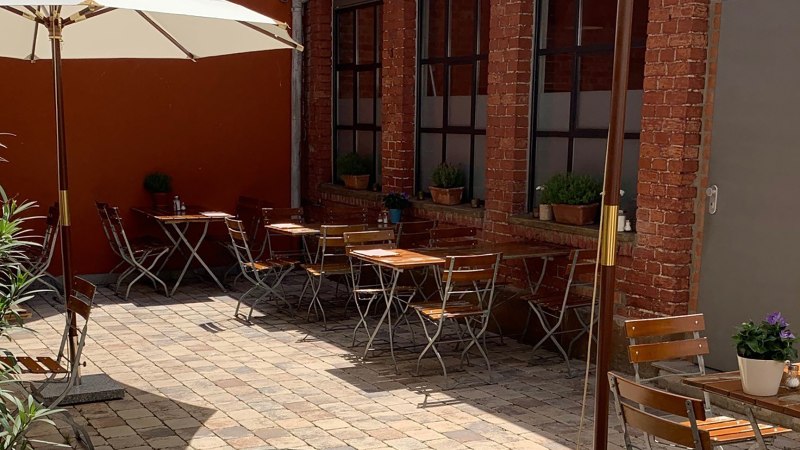 Cozy courtyard with wooden tables and chairs, parasol and plants. Red brick walls and paved floor create an inviting atmosphere., © Gustav Gastro GmbH