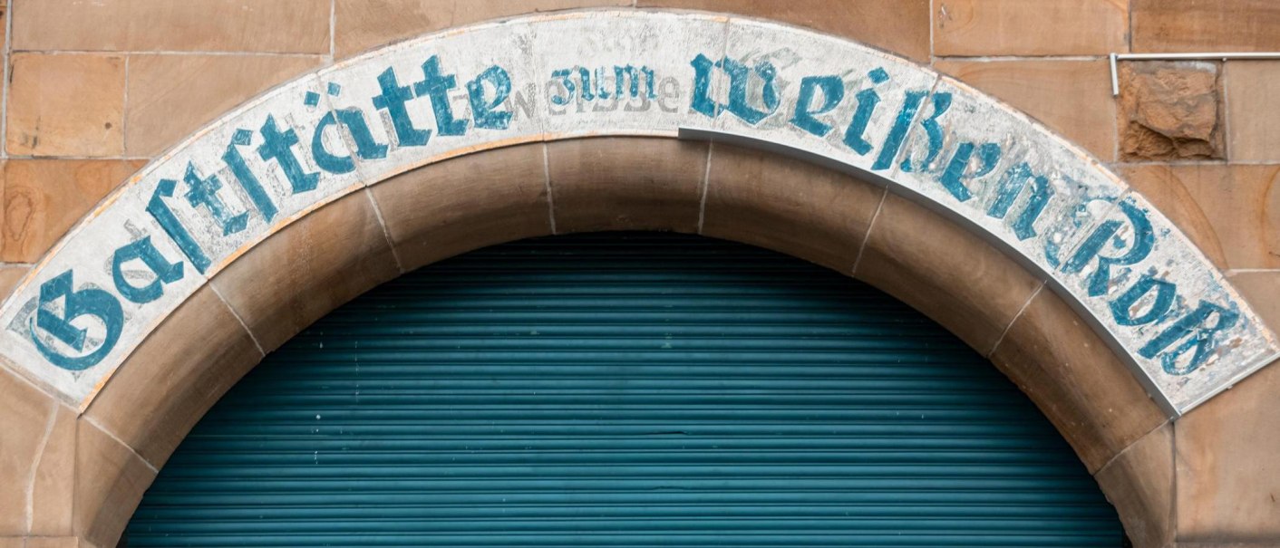Ein altes Schild &uuml;ber einem Torbogen tr&auml;gt die Aufschrift 'Gastst&auml;tte zum Wei&szlig;en Ro&szlig;'. Der Hintergrund ist ein geschlossenes, blaues Rolltor., &copy; Stuttgart-Marketing GmbH, Sarah Schmid
