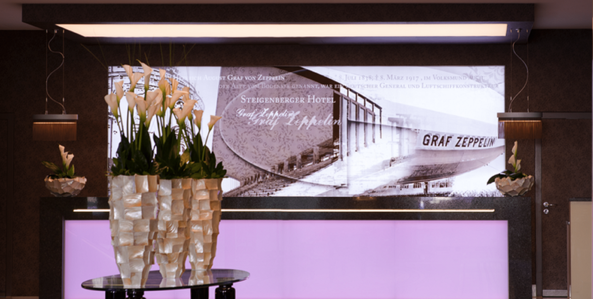 Elegant hotel reception with a table full of flowers. In the background a large picture of Graf Zeppelin and the lettering 'Steigenberger Hotel'., © Steigenberger Graf Zeppelin