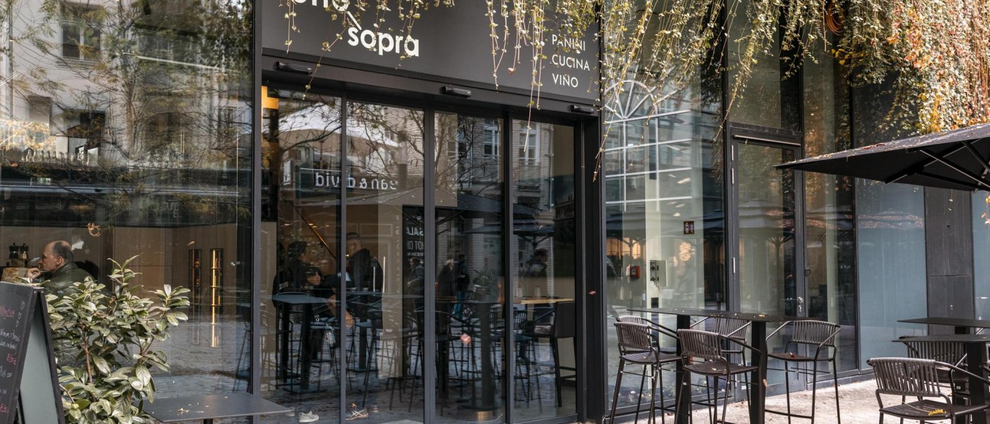 Eingang eines Cafés mit Glasfront und Außenbestuhlung. Pflanzen hängen von oben herab. Menschen sitzen drinnen. Schild mit 'sotto sopra' sichtbar., © SMG, Sarah Schmid