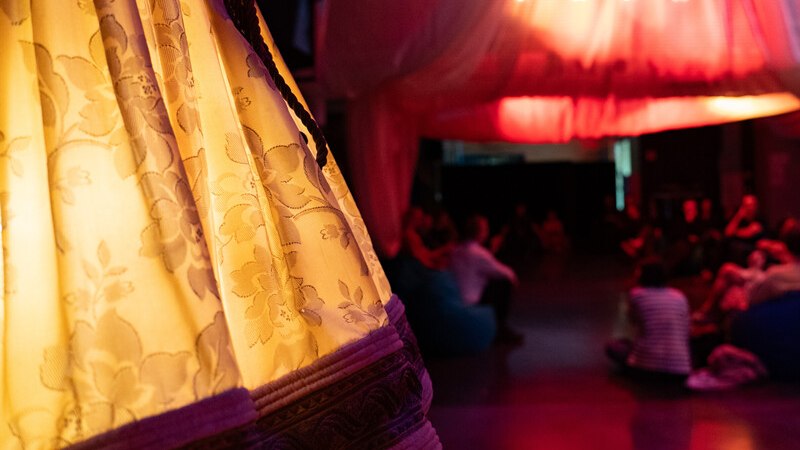 Ein Raum mit gedämpfter Beleuchtung, dekoriert mit Stoffen. Menschen sitzen entspannt auf Sitzsäcken. Im Vordergrund ein beleuchteter Vorhang., © Württembergische Staatstheater Stuttgart