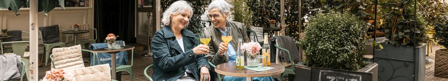 Zwei Frauen sitzen in einem gemütlichen Café im Freien, stoßen mit Getränken an und lachen. Ein Schild bietet Zimtschnecken an., © Stuttgart Marketing GmbH - Sarah Schmid