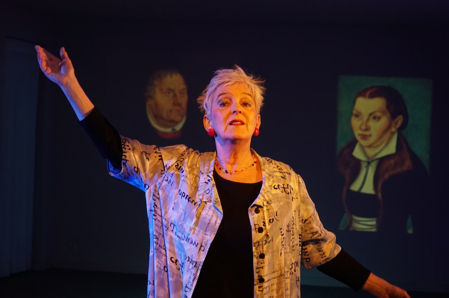 Eine Frau steht auf einer Bühne und gestikuliert. Im Hintergrund sind Projektionen von zwei historischen Porträts zu sehen., © Katja Ritter Eine Frau steht auf einer Bühne und gestikuliert. Im Hintergrund sind Projektionen von zwei historischen Porträts zu sehen., © Katja Ritter