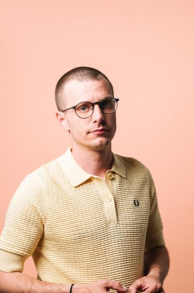 Ein Mann mit Brille und kurzem Haar trägt ein gelbes Polohemd vor einem pfirsichfarbenen Hintergrund., © Rosenau Kultur e.V.