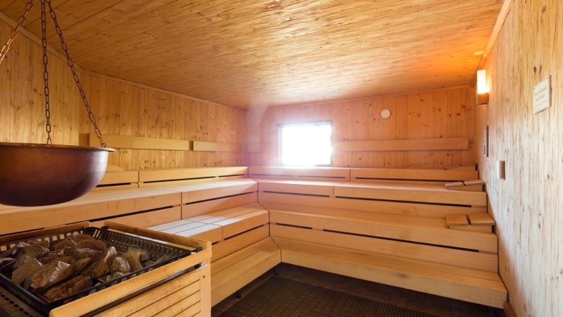 Holzverkleidete Sauna mit Bänken und einem Ofen mit Steinen, warmes Licht und kleines Fenster., © ARAMIS Hotel & Freizeit GmbH