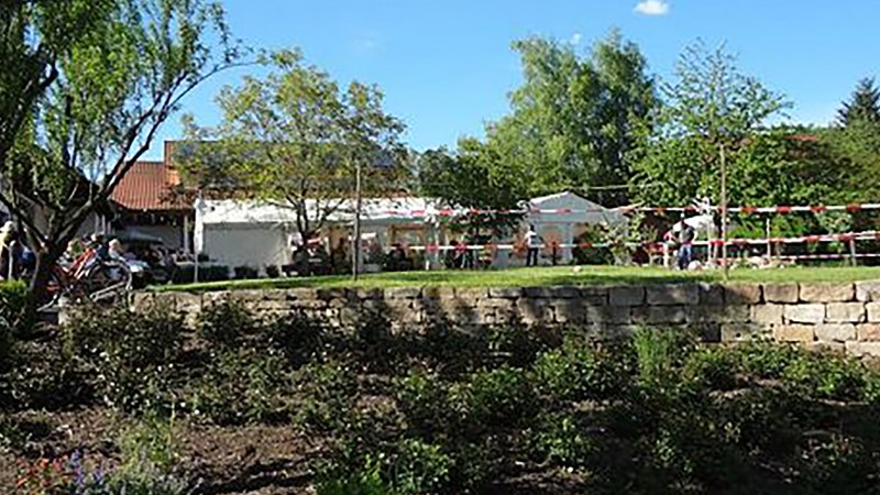 Ein Garten mit Bäumen und Menschen bei einem Fest. Im Hintergrund sind Zelte und ein Gebäude zu sehen. Der Himmel ist blau und sonnig., © Weingut Klopfer