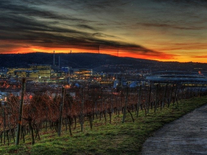 Evening view of a city with illuminated buildings and a stadium, surrounded by vineyards. The sky is dramatically colored red-orange., © Cool-Tours StattReisen Evening view of a city with illuminated buildings and a stadium, surrounded by vineyards. The sky is dramatically colored red-orange., © Cool-Tours StattReisen